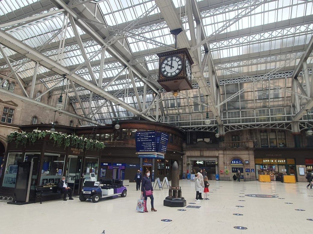 Old-fashioned clock in wooden casing with Roman Numerals hanging from a glass roof at Glasgow Central train station.