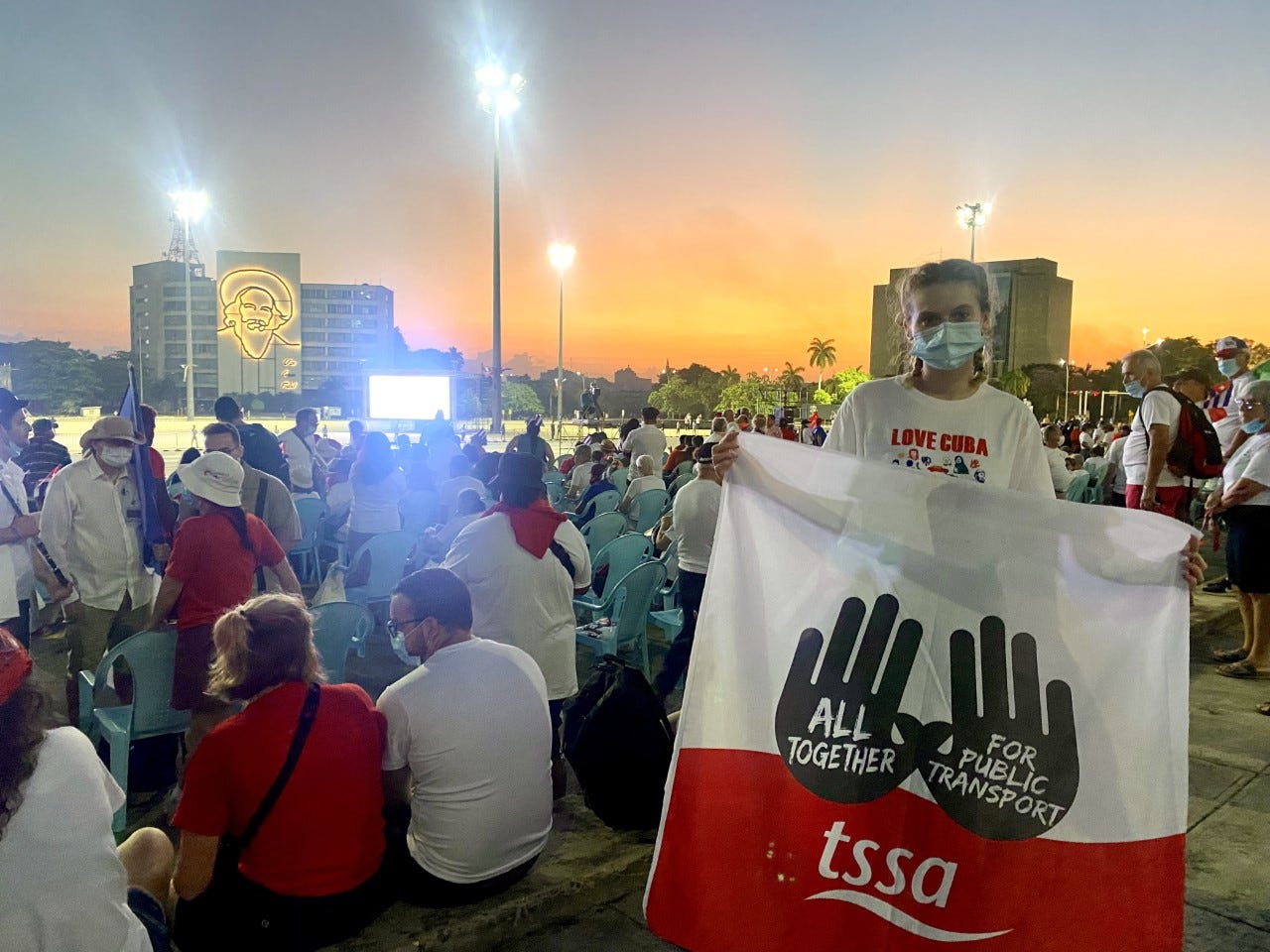 Maisie D stands holding a TSSA Banner in front of a large group of people wearing red and white t-shirts with their backs facing the camera. They are facing a screen and a building with an image projecte don it. Maisie has pigtails and is wearing a face mask.