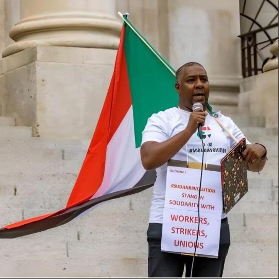 The image shows an image of Khalid Sidahmed, a well known Black trade unionist, standing speaking at a rally about standing in solidarity with Sudan whilst holding a flag of Sudan. He wears a sign around his neck that reads, 'Stand in Solidarity with WORKERS, STRIKERS, UNIONS.' 