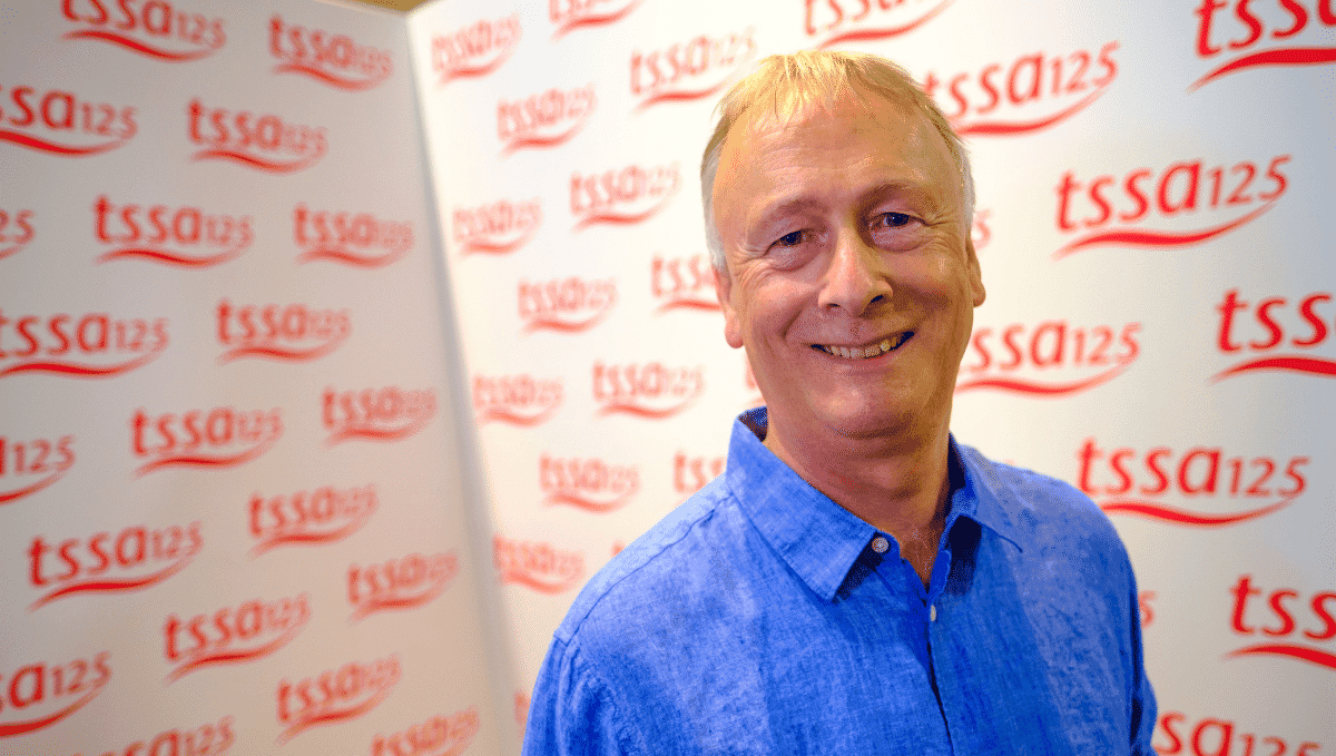 Steve Coe, an older white man, in bright blue shirt smiles at the camerain front of a white wall covered in tssa125 logos. 