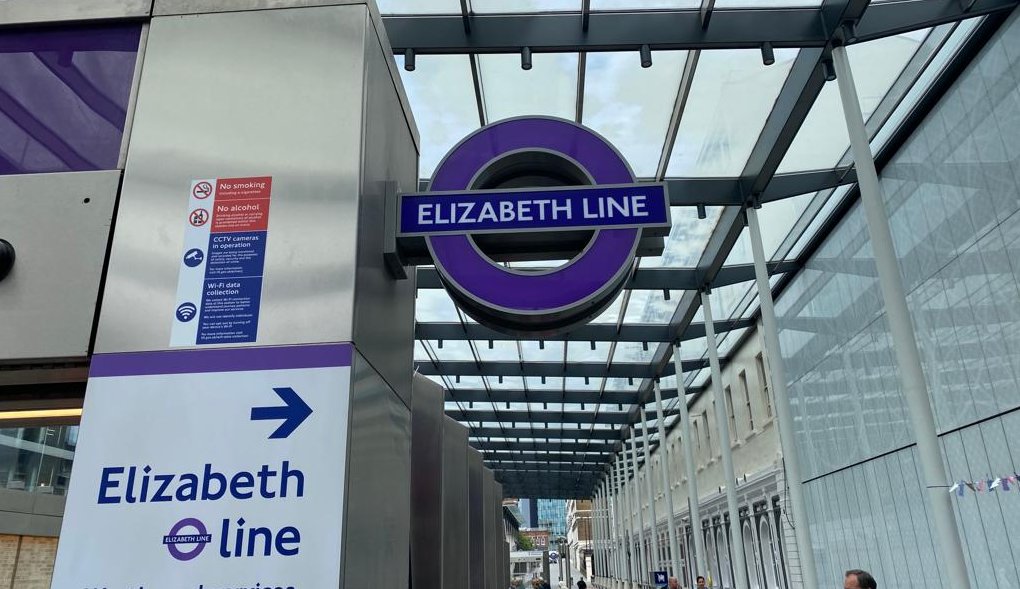 a shiny new corridor of chrome and glass. There is a purple Elizabeth Line rondel above the corridor and and a sign with an arrow pointing to the corridor which says Elizabeth Line.