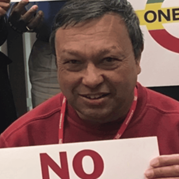 Anand Pillar, a middle-aged Asian man with greying hair in a red sweater. He is holding a white sign with the word "NO" on it in red.
