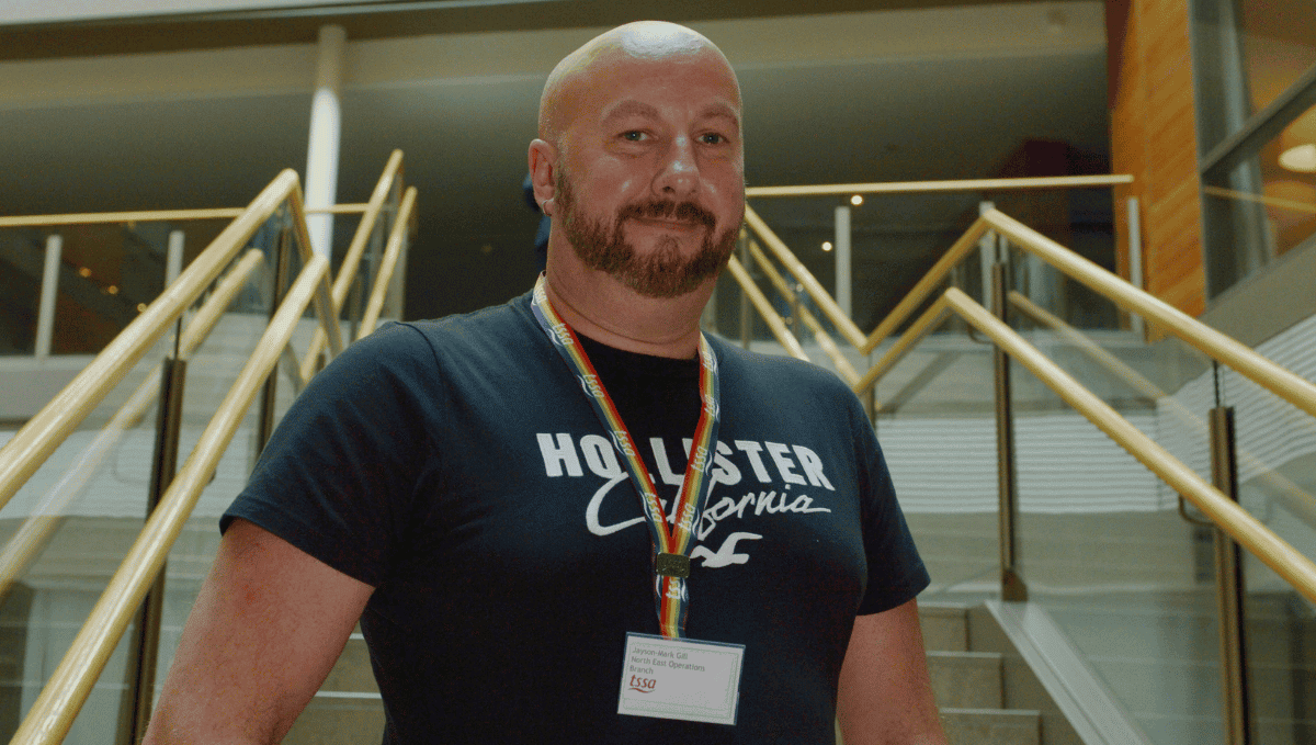 EC member Jayson-Mark Gill, a bald, white man, with a short beard wearing a black T-shirt and a rainbow lanyard stands on a flight of stairs looking at the camera.