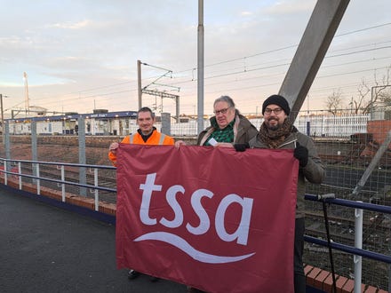 Three TSSA activists holding a huge TSSA banner. They are white men of various ages and sizes. In the background is Airdrie train station and ticket office. 