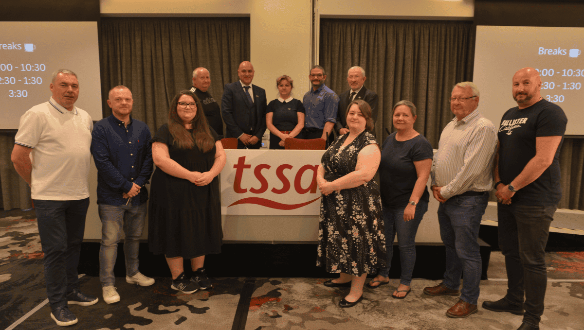 Current and former members of the TSSA Executive Committee from different genders, ages and races stand with TSSA Interim General Secretary Peter Pendle and a podium with a TSSA flag. 