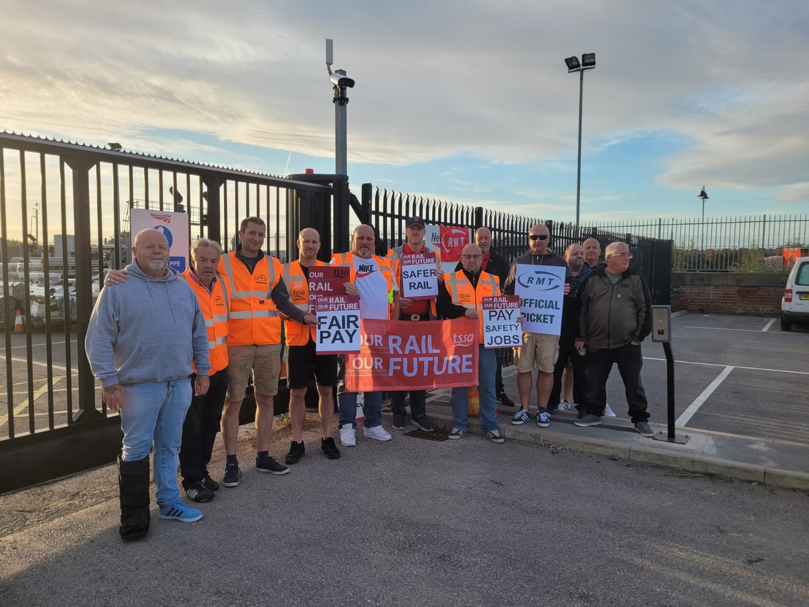 Image shows 10 union members on a picket line in hi-vis jackets on a sunny day holding a red 'our rail our future' banner