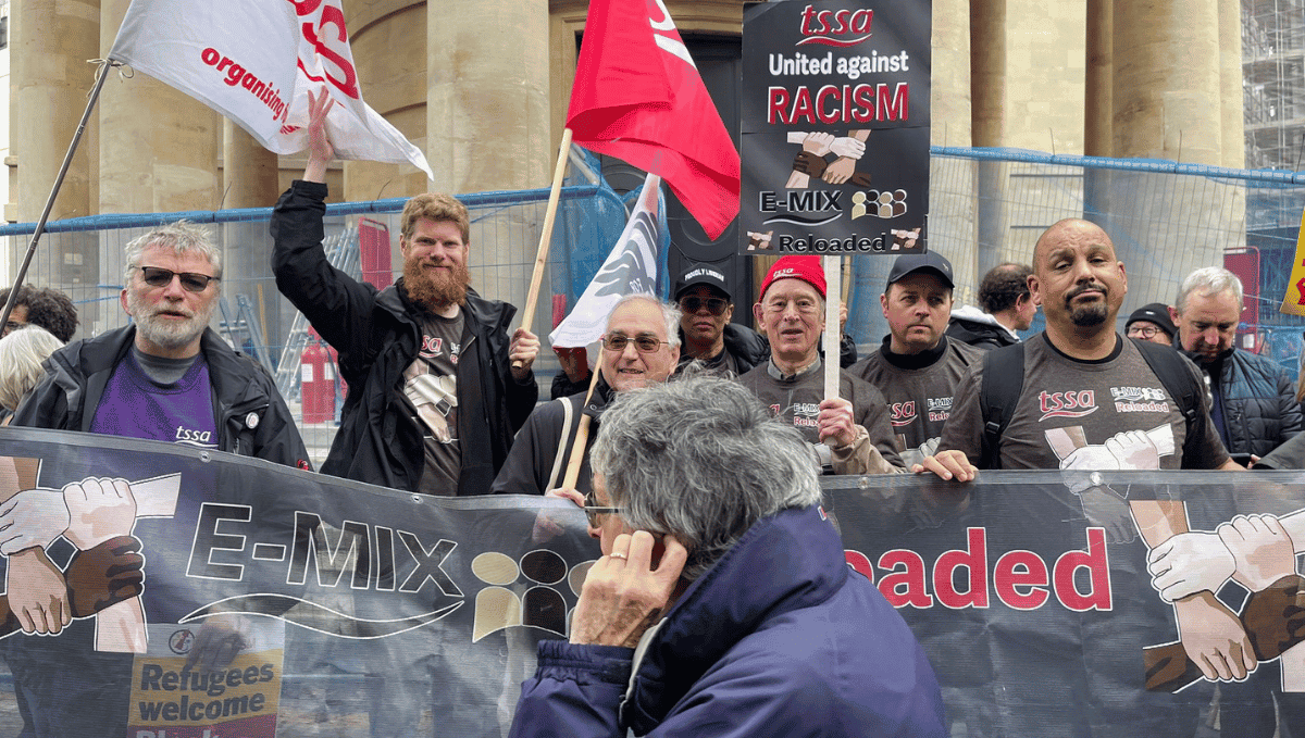 A group of TSSA activists of varying ages and races wearing TSSA E-mix T-shirts, waving TSSA flags and holding up a big TSSA E-Mix Reloaded banner.
