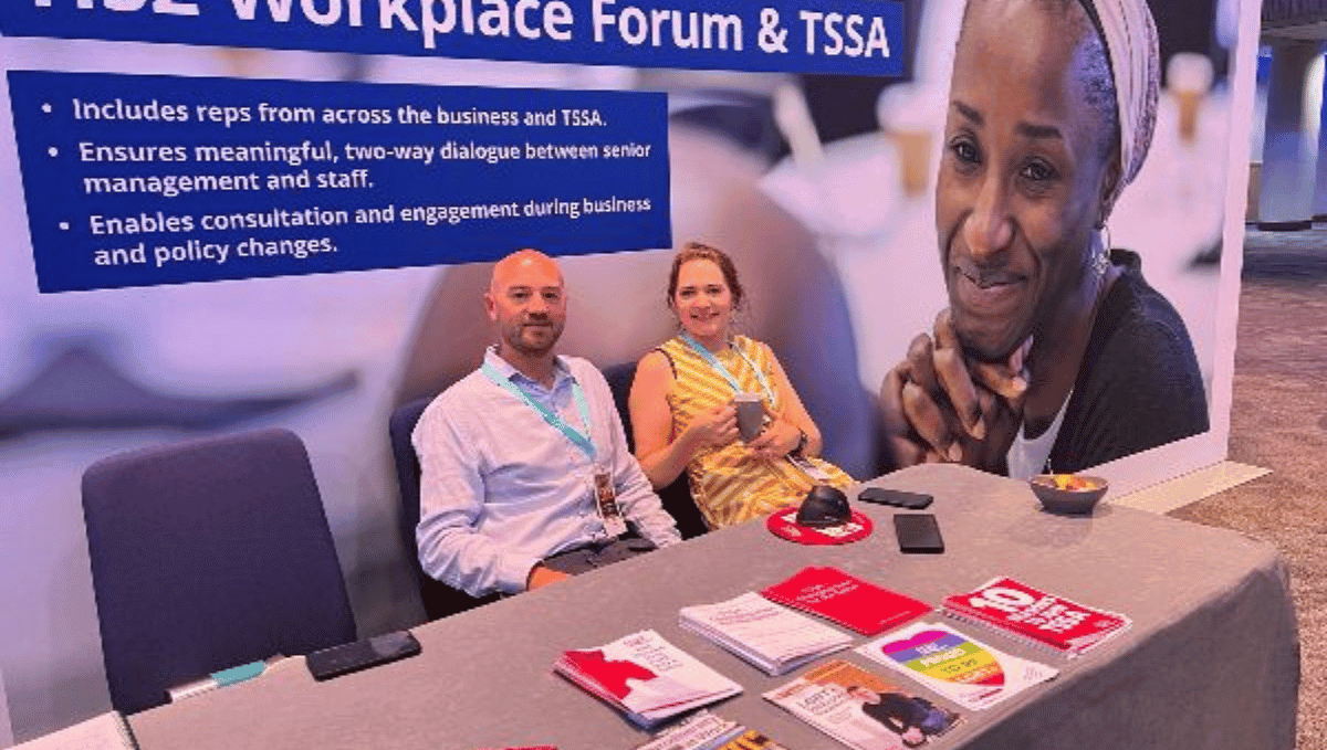 Two people sitting in front of a huge pop up banner with a desk in front of them. The desk has TSSA recruitment materials on it. The man is wearing a purple shirt and blue lanyard, the woman is wearing a yellow dress and holding a cup. The pop up banner says "HS2 Workplace forum and TSSA" and has a picture of a black woman on it.