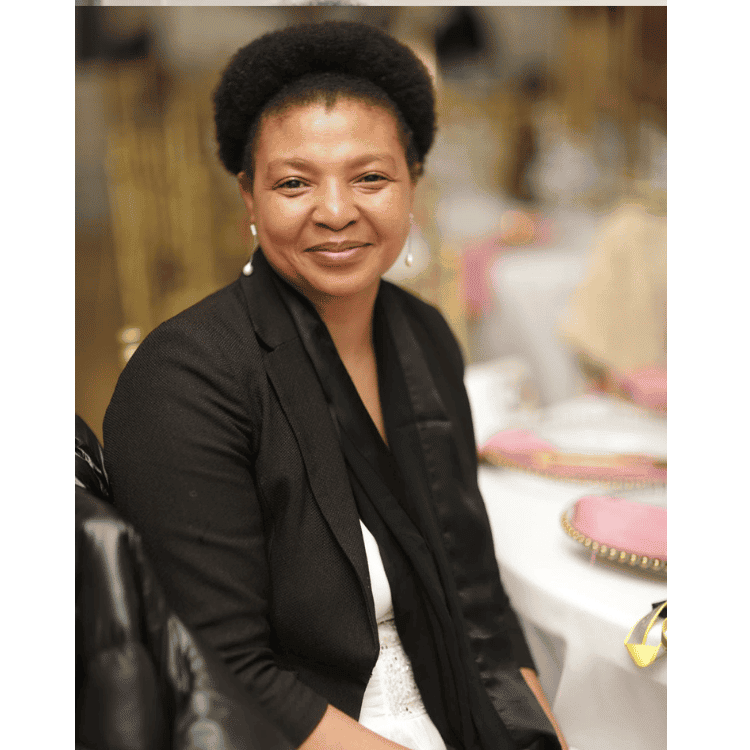 Mary Sithole, a black woman with natural hair smiling at the camera. She is seated at a circular table with a white tablecloth and pink and gold tablewear. Mary is wearing a white dress with lace trim, and a black jacket with a decorative black scarf, and pearl drop earrings.