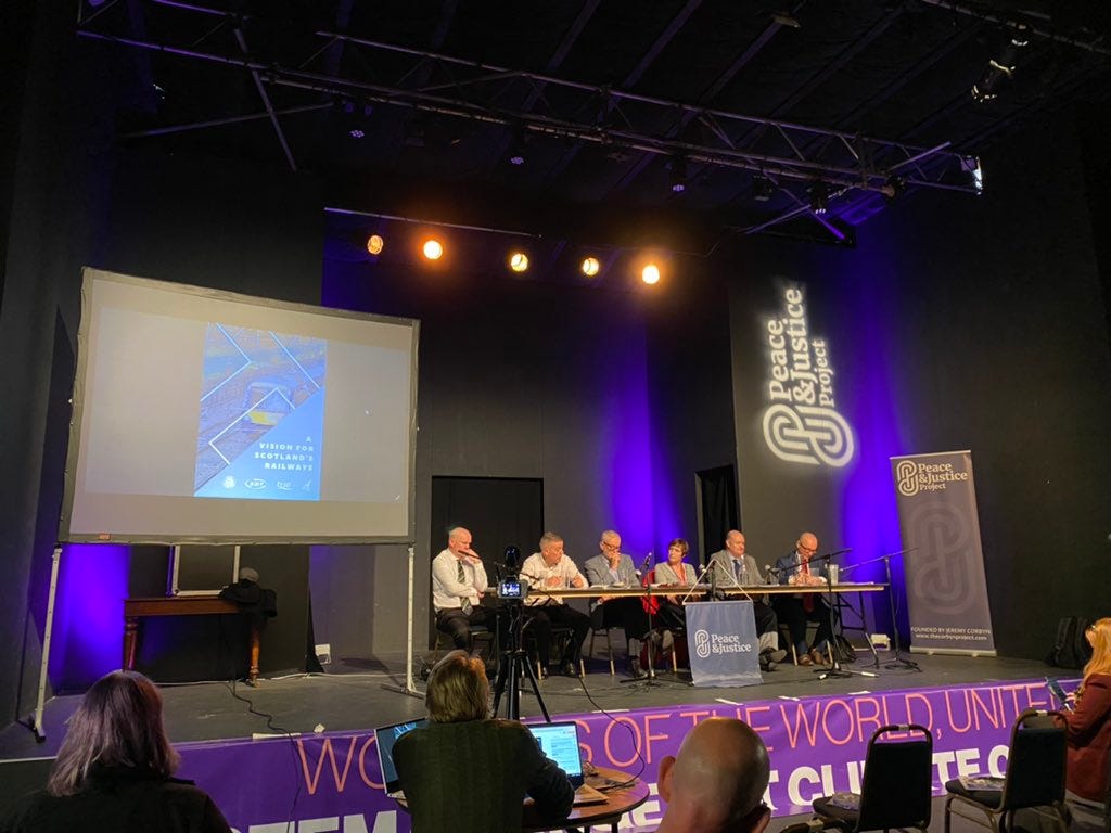 4 rail union leaders, Jeremy Corbyn and Roz Foyer at a table on a stage at COP26. A Peace and Justice Project banner to one side and a screen showing the report "A Vision for Scotland's Railway" is above them. Photo credit Niall Christie