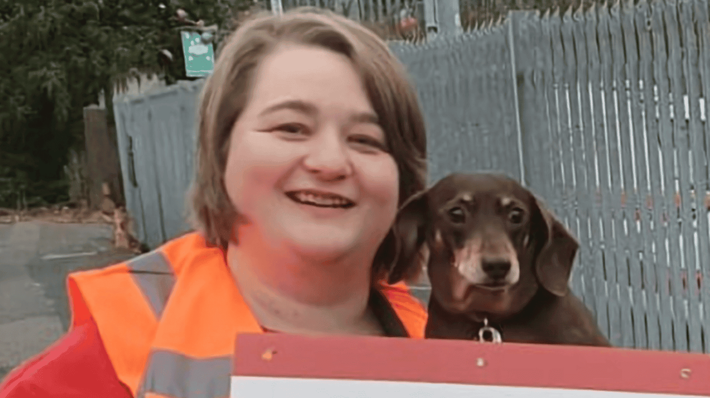 Melissa Heywood, a white woman with chin-length brown hair, wearing an orange hi-vis vest over a red t-shirt. She is holding a little brown dog with floppy ears at her shoulder-height.