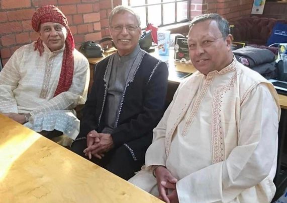 Three asian men in traditional clothing. The man in the middle wears a dark robe with embroidery on the shoulders, pockets and edges over a grey shirt. The man on the left wears a robe with pale green and grey stripes trimmed with gold embroidery. He has a red headscarf with a gold pattern which is wrapped around his head and hangs over one shoulder. The man on the right is in white with red embroidery on the edges. They are all smiling.