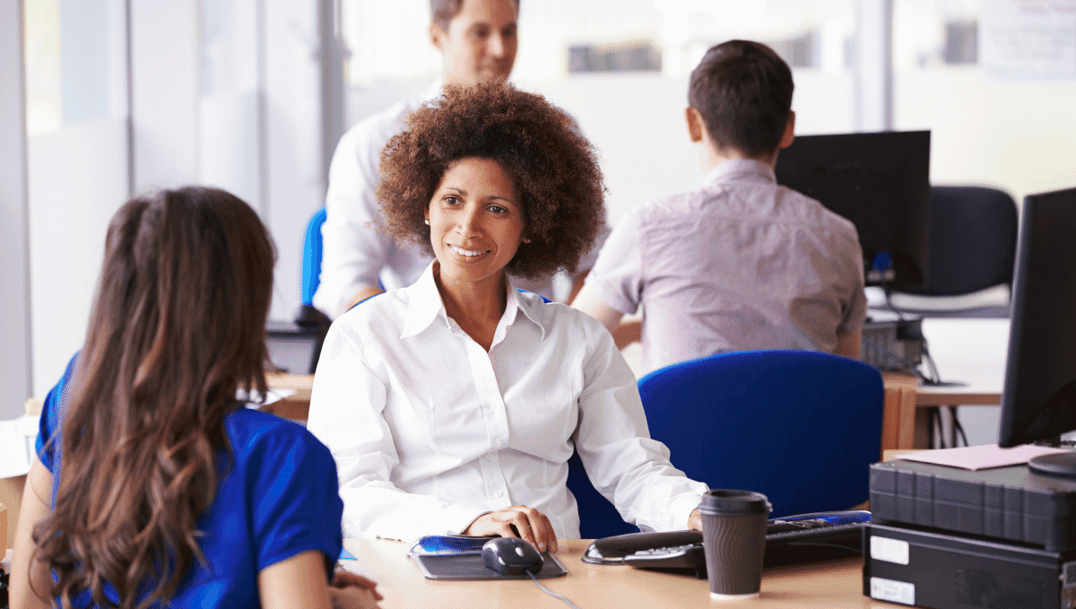 Two women sat at a desk talking with two men at a desk in the background