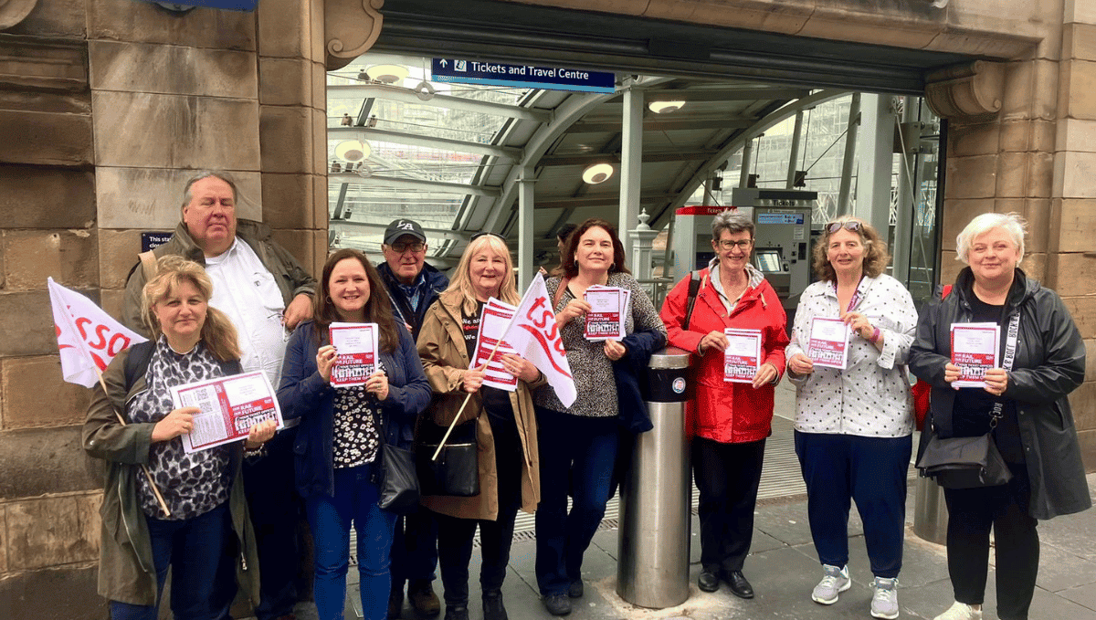 A row of 9 people of various ages, men and women, holding TSSA flags and leaflets opposing ticket office closures standing outside the entrance to Edinburgh Waverley.