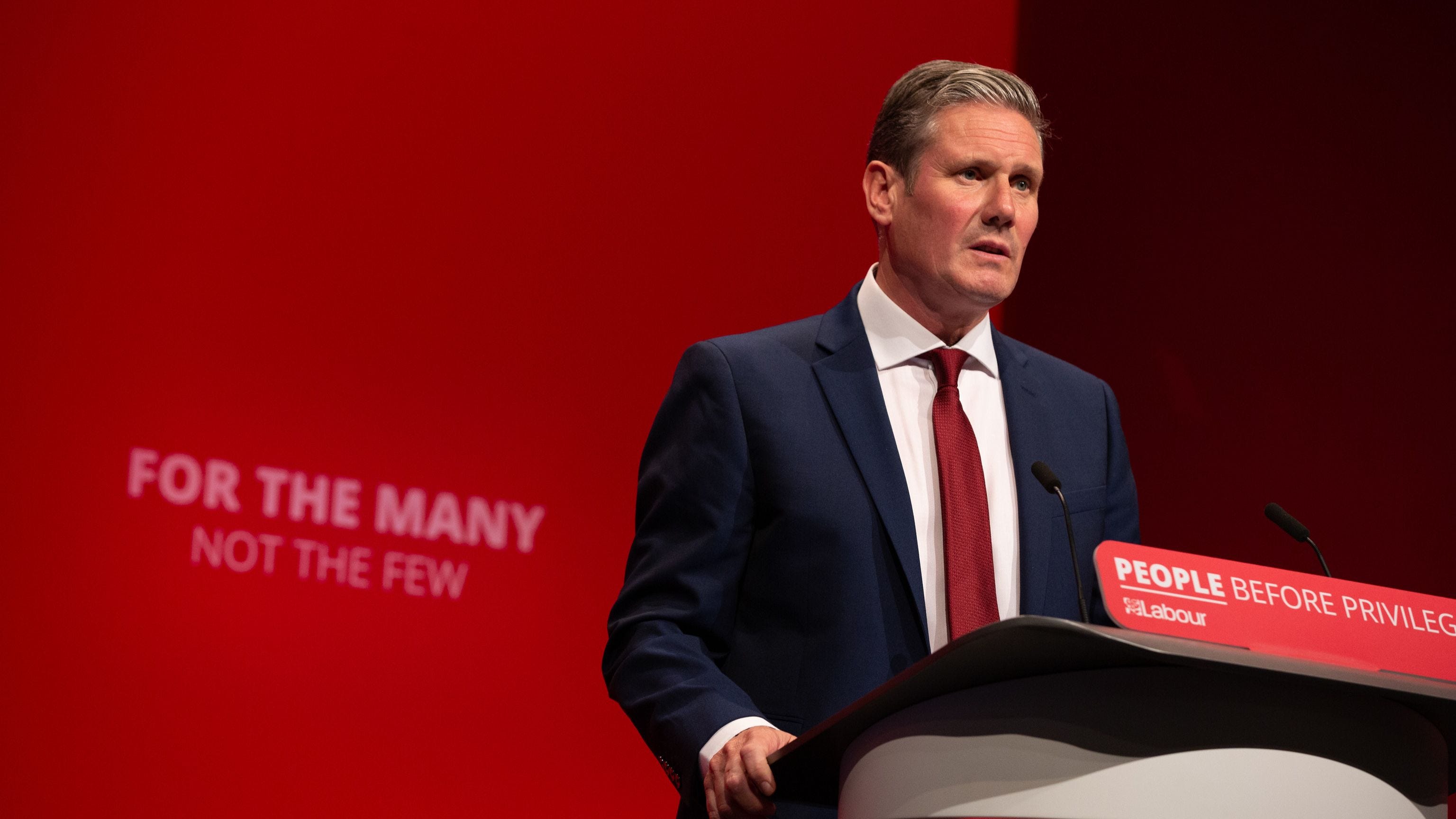 Keir Starmer speaking at the podium at Labour Party conference with red background showing slogan: For The Many Not The Few
