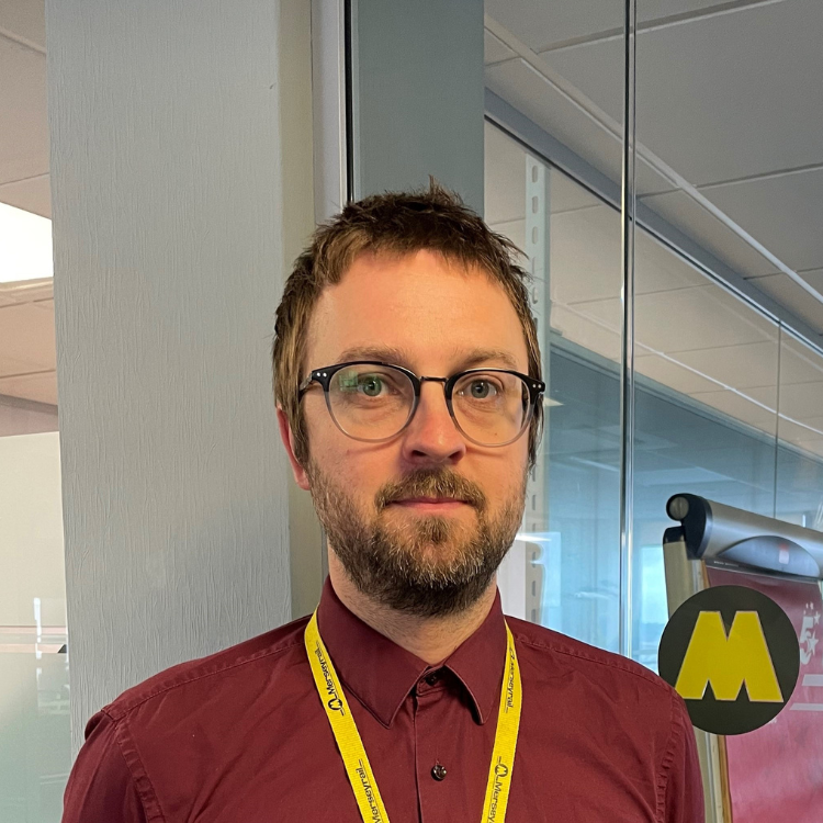 Ian a white man with brown hair and beard, blue eyes, and black framed glasses. He is wearing a maroon shirt with a yellow lanyard. Behind him a yellow Merseyrail 