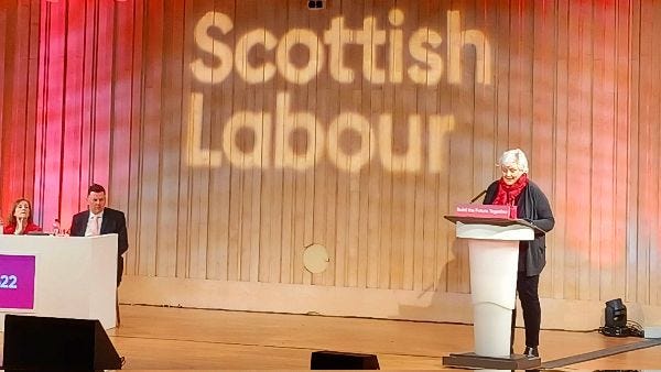 Image shows Katrina Faccenda, a woman with white hair, black trousers, a grey cardigan and a red scarf. She is standing speaking at a podium. Behind her there are red and yellow curtains with the words "Scottish Labour" on them. To the left of the picture is a man and a woman sitting at a desk.
