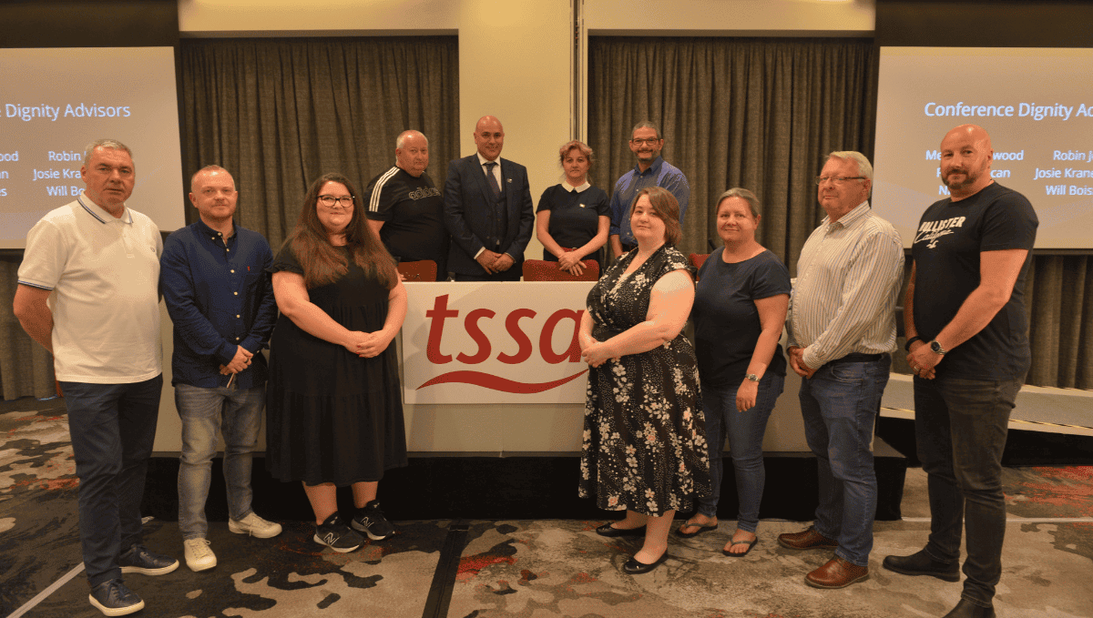 A group of male and female executive committee members of various ages, gathered around a podium with a TSSA flag.