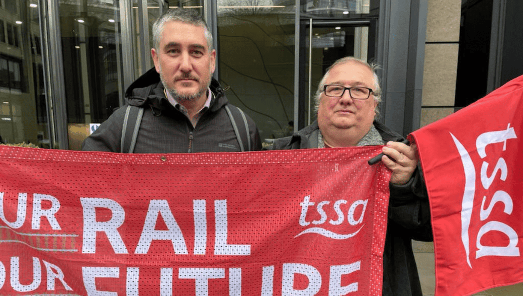 Luke Chester and Mick Carney holding TSSA flags outside Rail Delivery Group 