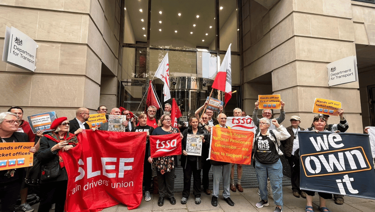 A large crowd of protestors of various races, ages and genders including TSSA's Mel Taylor on the left and Bonnie Craven in the centre. The protestors are holding banners from TSSA, ASLEF and We Own It.  They are standing outside the Department for Transport, a large concrete building.