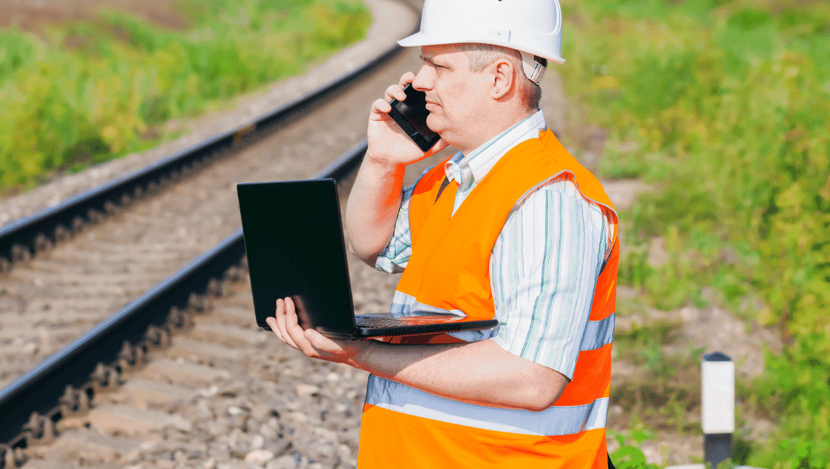 Rail worker in orange high vis trackside with laptop
