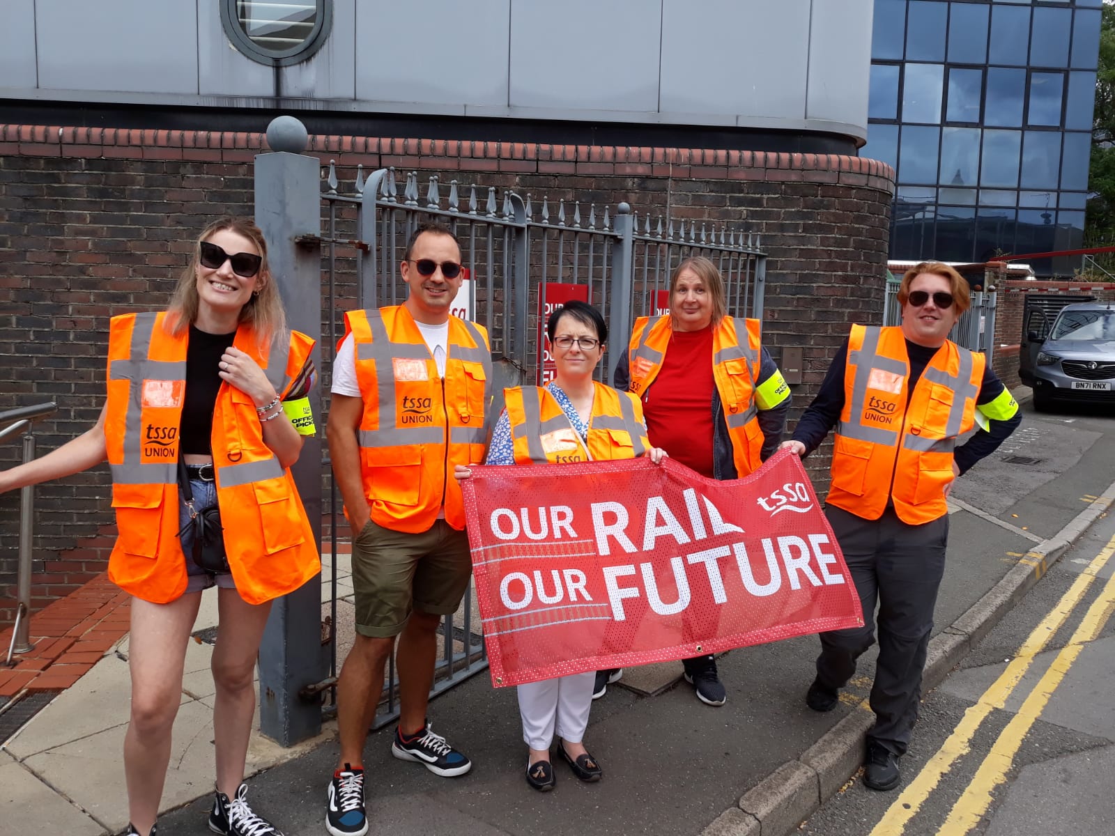 Photo shows five TSSA members on picket line on a sunny day in Swindon, holding red 'Our Rail Our Future' Banner