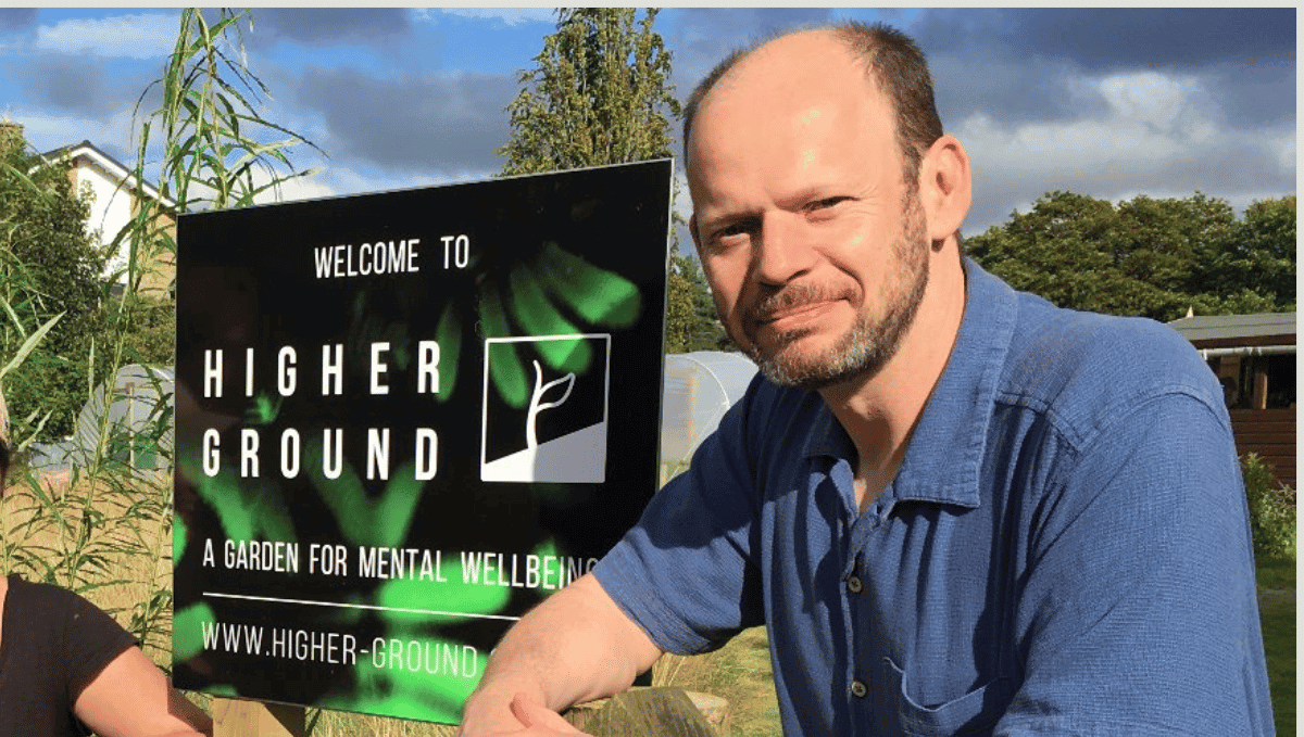 Jamie Driscoll, a white, balding man, lightly tanned, with a greying beard and moustache and prominent ears. He is wearing a short sleeved, open-necked denim blue shrt, and seated beside a sign saying "Welcome to Higher Ground. A garden for mental wellbing".