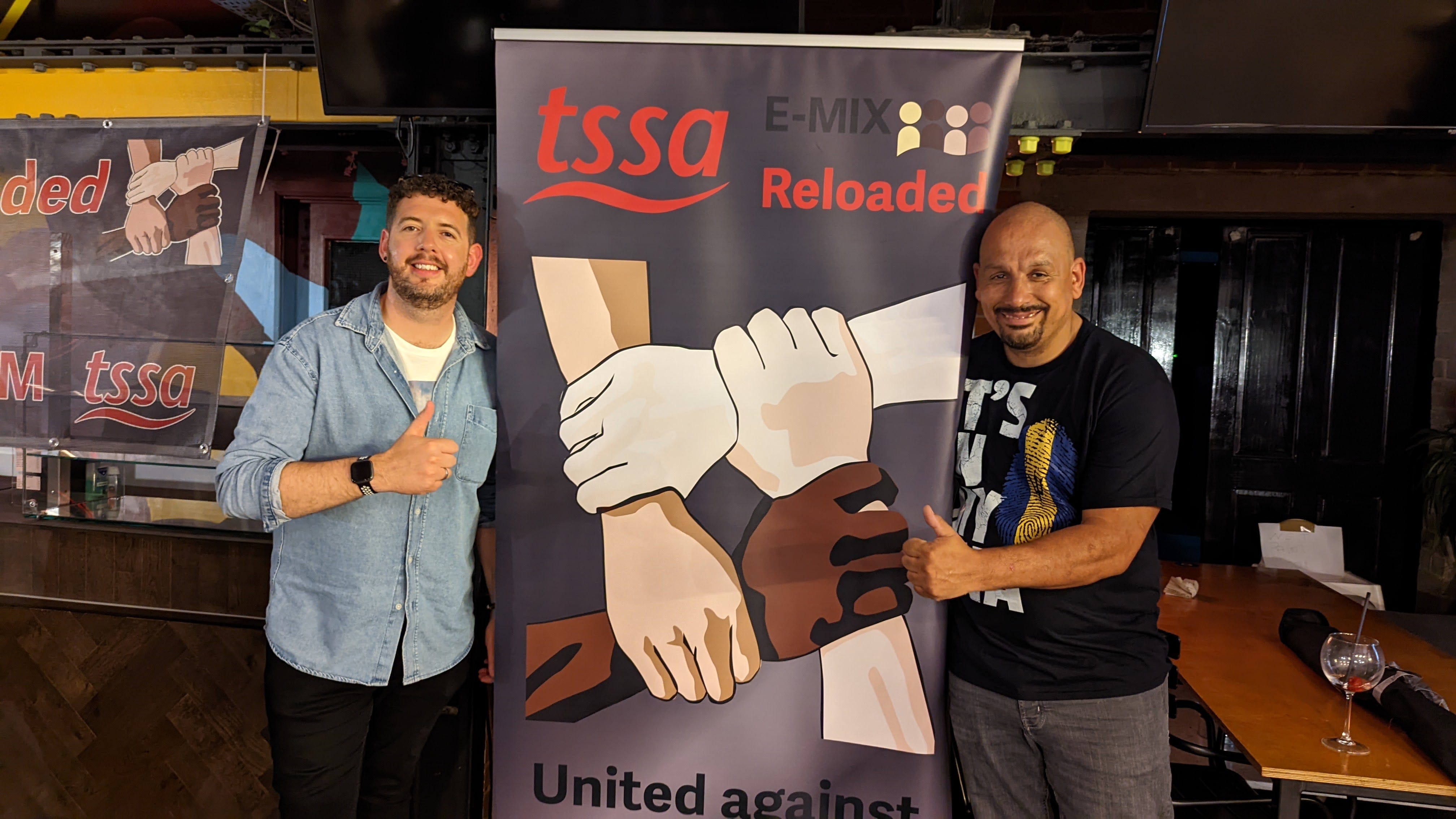 A white man and a black man stand either side of a pop up banner which reads "TSSA E-MIX Reloaded: united against racism. The white man is wearing a blue shirt with a white shirt underneath and the black man wears a black t-shirt. The Banner has four hands in 4 different skin colours clasping each other's wrists in a show of unity.
