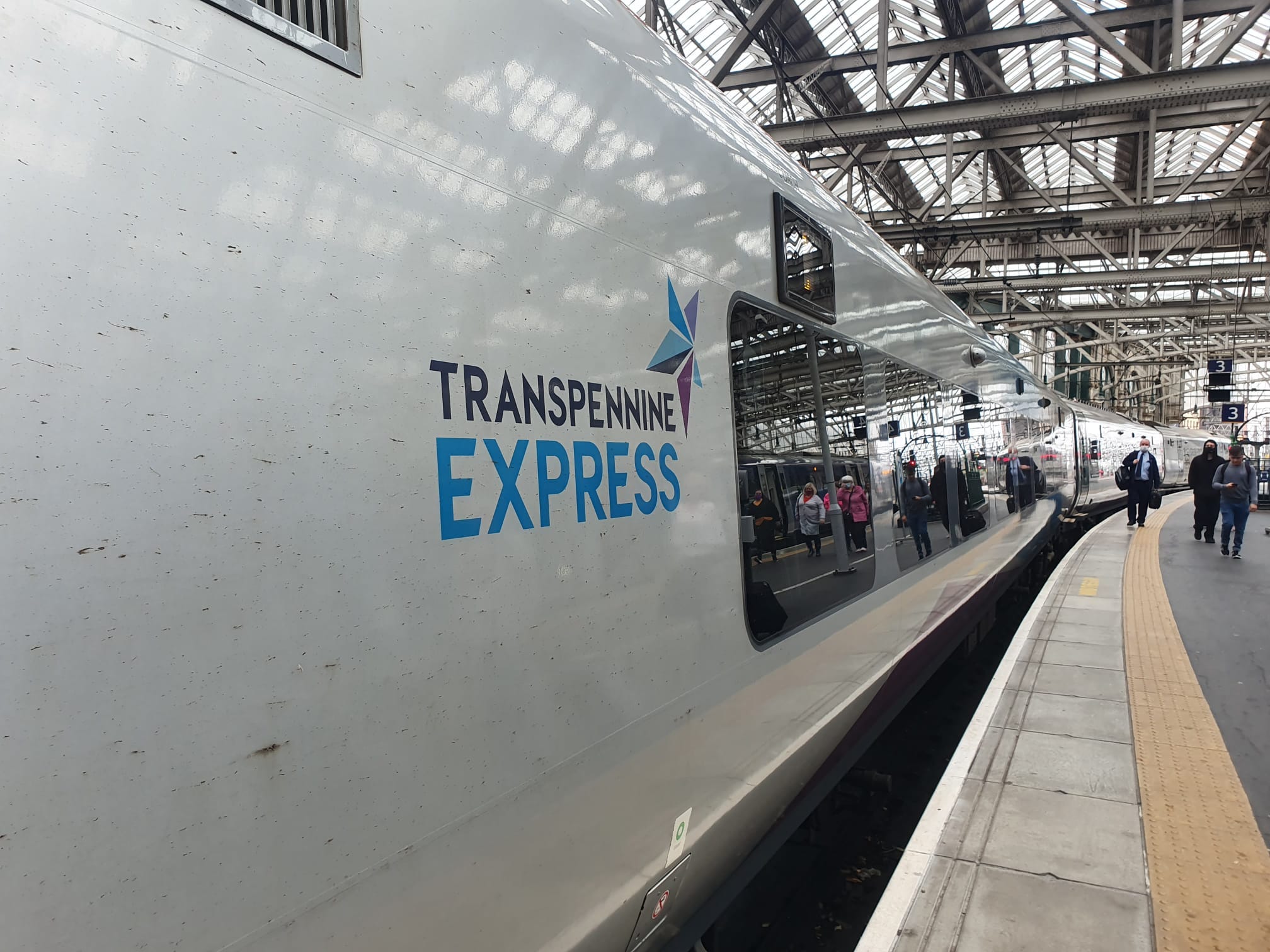 View along the side of a white train in a station, with the words "Transpennine Express" and a blue and purple star on the side. 