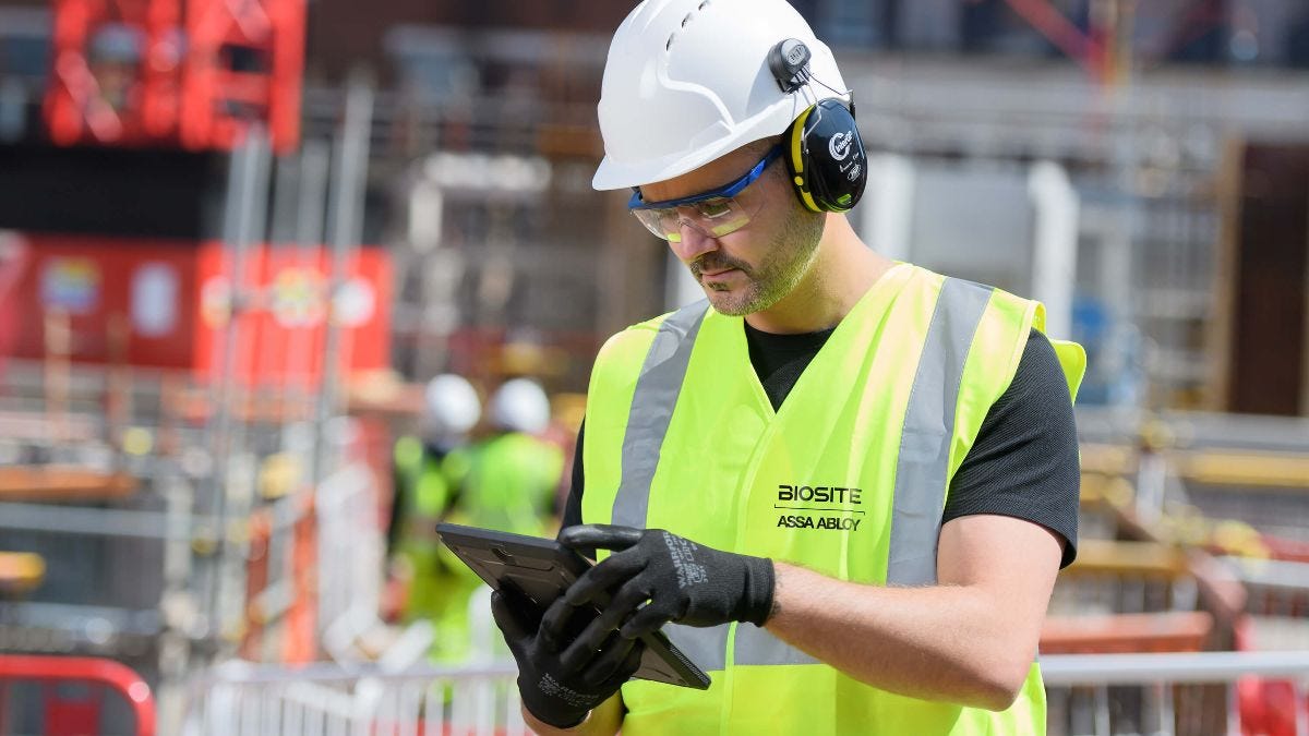Health and safety inspector wearing yellow vest, safety specs, ear defenders, gloves and hard hat using tablet.