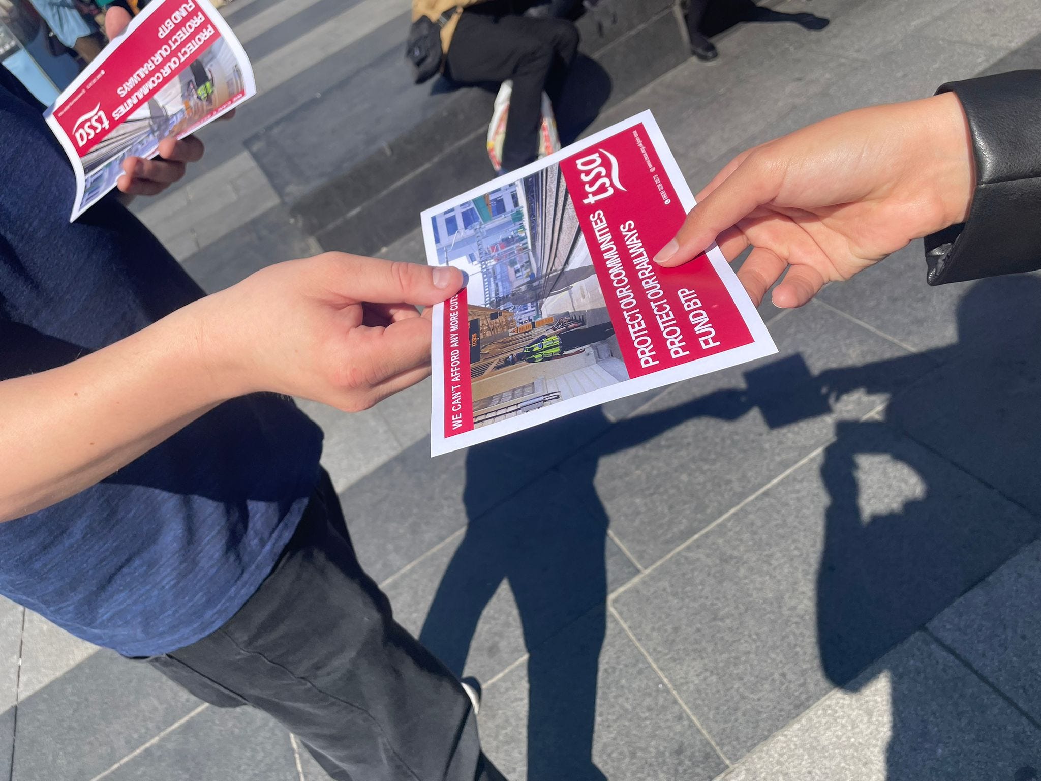One person hands one of TSSA's leaflets to another person. We can see their hands but not their bodies or faces.