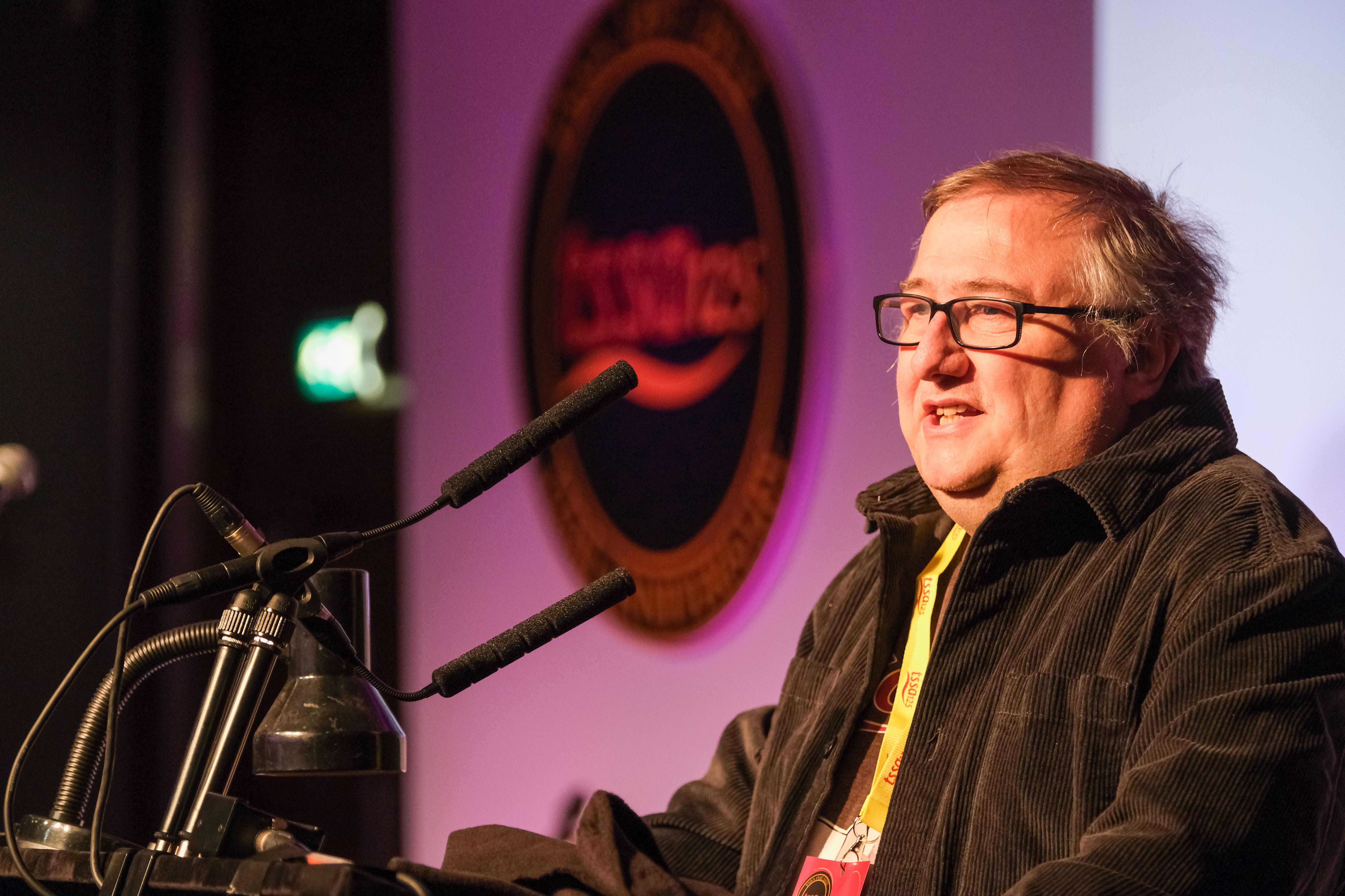 Mick Carney a white man with glasses, wearing a brown fleece and a yellow lanyard speaks at a rostrum in front of the TSSA125 logo 