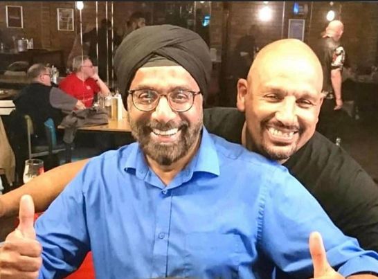 An Asian man in a blue shirt and a black turban gives a thumbs up to the camera and a black man in a black t-shir hugs him from behind. They are both grinning happily.