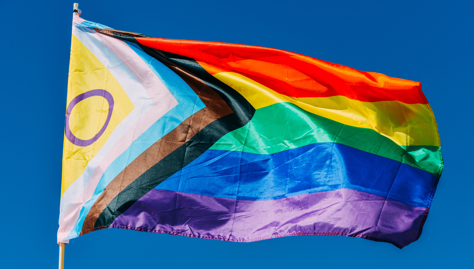 An inclusive pride flag against a bright blue sky. The flag features a rainbow, red, yellow, green, blue and purple top to bottom, with a chevron on the right of black, brown, powder blue, pale pink and white stripes. Inside the chevron is a yellow triangle with a purple circle. 
 