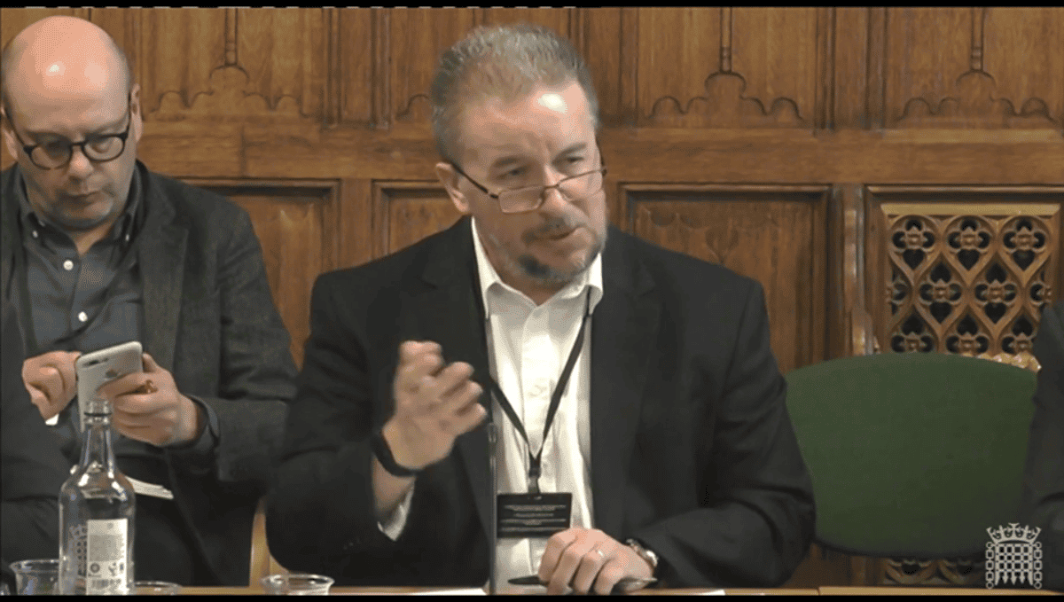 Rob Jenks, a white man with grey hair and short beard, giving evidence in a Westminster Parliamentary select committee room. The wall is wood pannelled. Rob is wearing a black suit jacket, white shirt and glasses. His hand is in the air.   