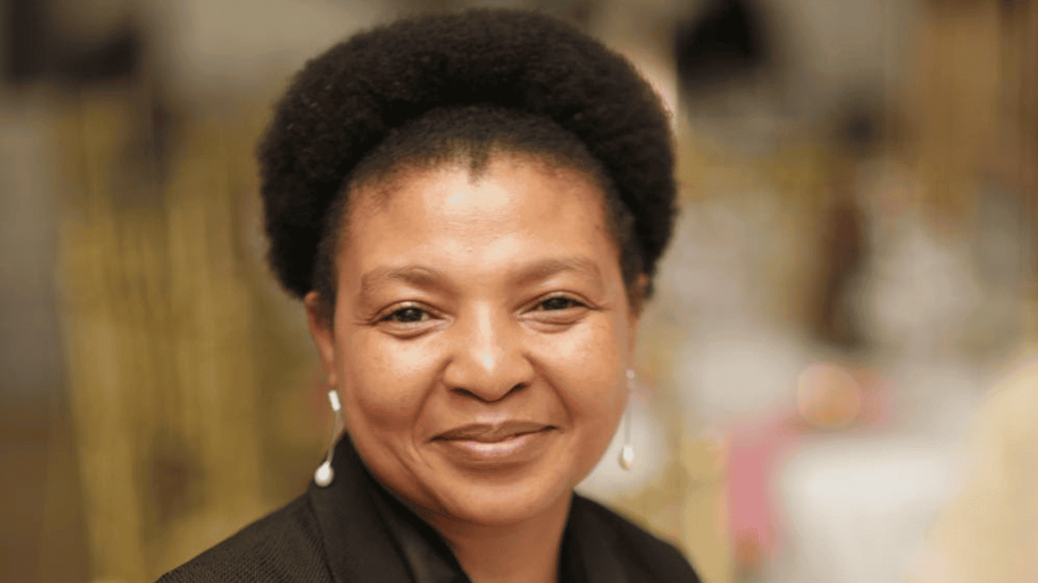 Mary Sithole, a black woman with natural hair smiling at the camera. She is wearing a smart black jacket and pearl drop earrings.