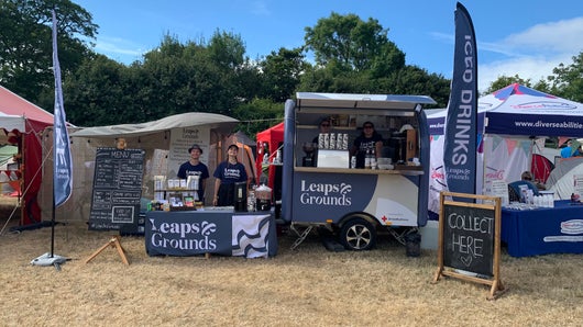 The Leaps & Grounds Coffee cart at Bestival. Two people stand in the cart while two more people stand beside the cart at a coffee station. All are wearing branded Leaps and grounds t shirts and smiling at the camera. There is also a sign in front of the cart that has 'collect here' written on it in chalk.
