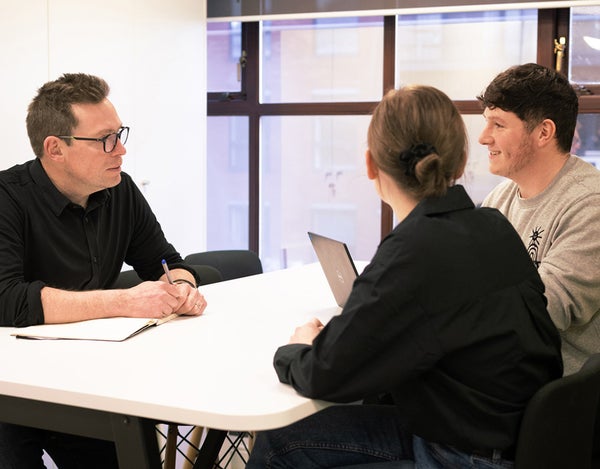 Three 26 employees sat around a table with a laptop and a notebook, talking.