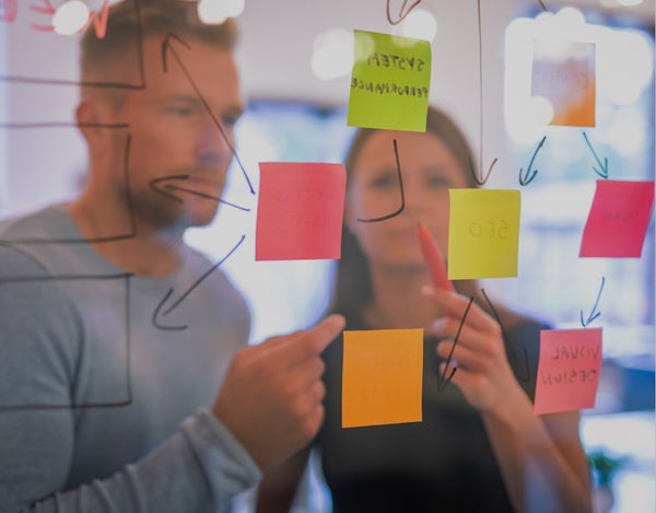 Two people standing in front of a window on which there are post-it notes, and discussing, while pointing at the post-its. One of the persons is holding a pen.