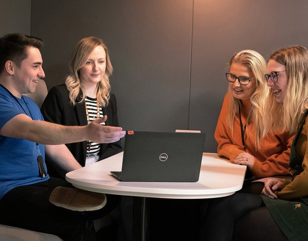 Four 26 employees sat around a table, talking, smiling and laughing. They are all looking at a laptop, on the table, and one of the employees is pointing at it.