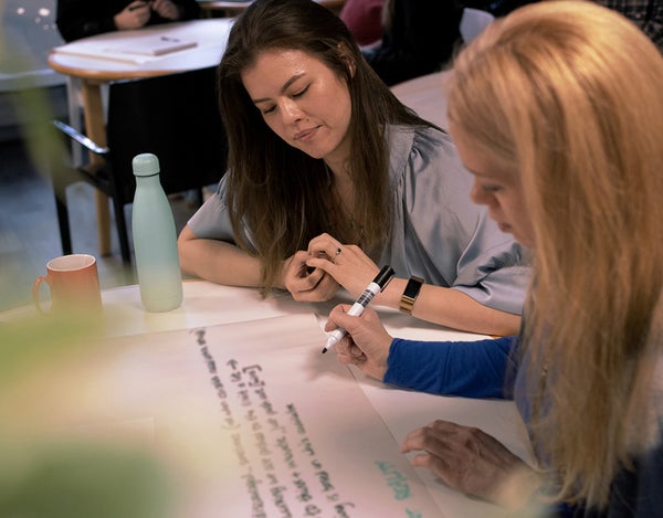 Two 26 employees sat at a table, one is holding a pen and writing on a large sheet of paper. There is a travel mug and a mug on the table.