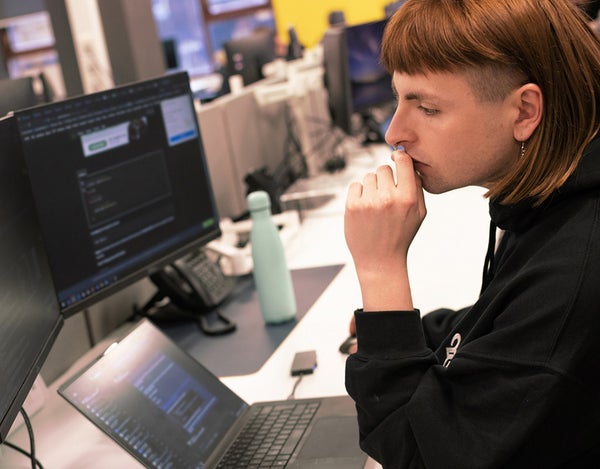 One 26 employees sat at a desk and looking at their computer screen, there is a laptop on the desk and they are looking at their extra monitor. There is a travel mug on the desk, and a office phone.