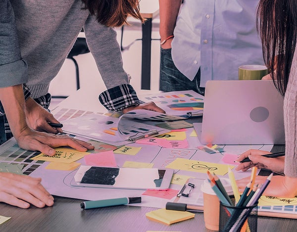 Four people standing around a table crowded with papers, post-its, a marker, and a full pencil holder.