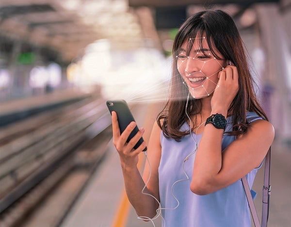 A person standing on a train platform, looking at their phone, smiling and holding headphones to their ear. Lines come out of the phone screen and surround the person's face.