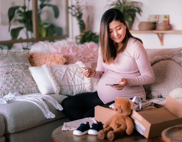 A pregnant person sat on a sofa and looking at their phone, surrounded by cushions, parcels and baby items.