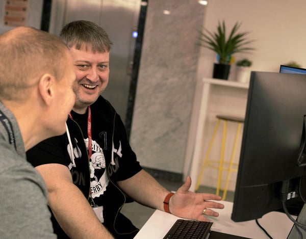 Two 26 employees sat at a computer desk and one of them is laughing.