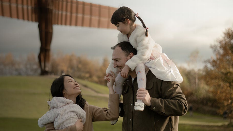 Family of 4 walking smiling at each other. Mother holding a baby and father holding a little girl. Angel of the North in the background. 