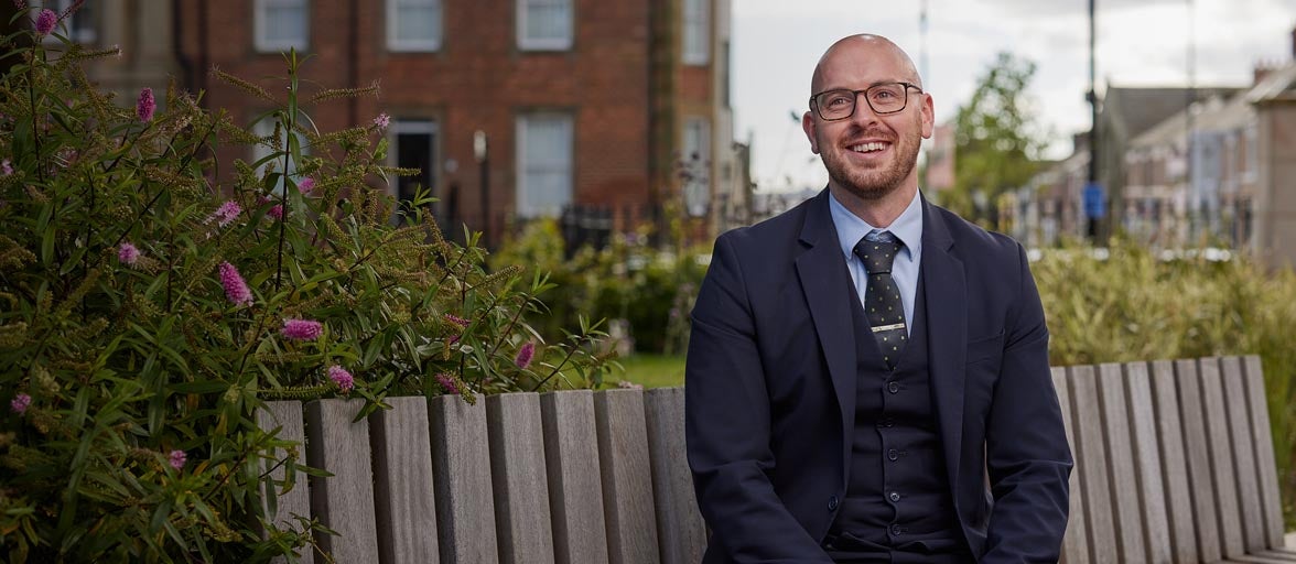 North Shields Branch Manager, Conor. Sat on a park bench smiling, with flowers in the background and a view of North Shields town. 