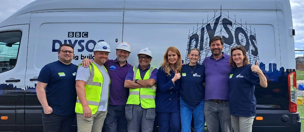 A group of Newcastle Building Society colleagues stood with members of the DIY SOS team, including Nick Knowles, beside a DIY SOS van. 