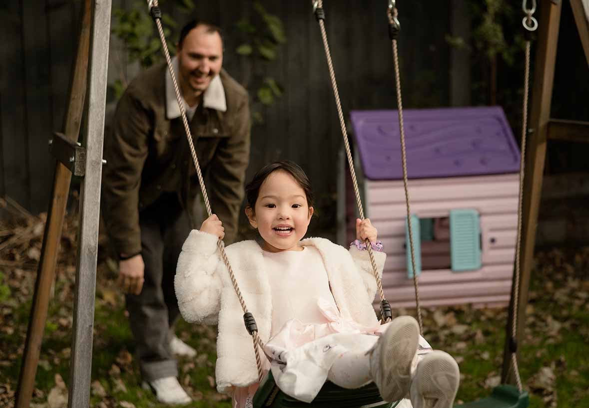 A Dad and his daughter outside in their garden, the Dad is pushing her on the swings. A play house can be seen in the background. 